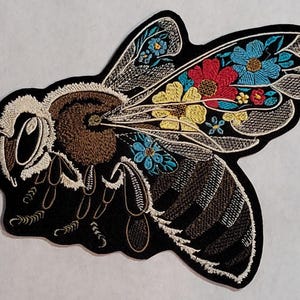 May include: An embroidered bee patch featuring a black body with brown fuzzy texture, white outlines, and wings decorated with vibrant red, yellow, and blue floral patterns. The insect shape is detailed with stitched segments and antennae.
