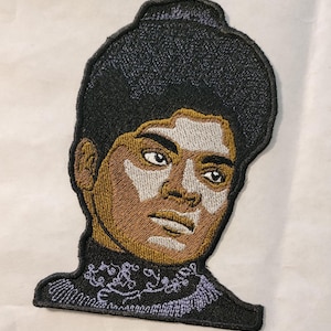 May include: Embroidered patch featuring a portrait of a person with dark hair, brown skin, and white makeup. The person is wearing a dark purple and black outfit with a decorative collar.