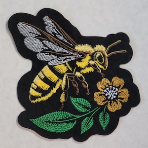 May include: An embroidered patch on a black felt background featuring a yellow and black bee with white wings hovering over a golden-yellow flower with green leaves. The design uses detailed thread work to depict the insect and floral elements.
