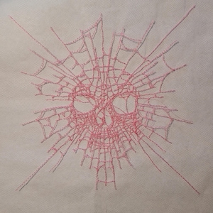 May include: Pink embroidered skull design with a spiderweb pattern. The skull's eye sockets and nose are visible within the web. The embroidery is on a light beige fabric.