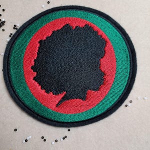 May include: Embroidered patch featuring a black silhouette of a person's profile within a red and green circular design. The patch has a black border and is likely for clothing or accessories.