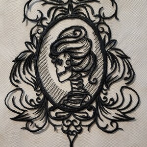 May include: Black embroidered design of a skeleton in a decorative frame. The skeleton is wearing a long dress and has a crown of flowers on its head.
