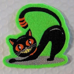 May include: An embroidered patch on a bright green felt backing featuring a black cat with orange eyes, an orange collar, and a wide yellow grin. The cat is arched in a stretching pose with a black and orange striped tail and stands on a silver base.