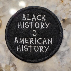 May include: A round, black patch with white embroidered text. The text reads "BLACK HISTORY IS AMERICAN HISTORY". The patch has a stitched border and is likely designed to be sewn or ironed onto fabric. The patch is a statement piece.