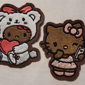 May include: Two embroidered patches featuring a brown cartoon cat. One cat is wearing a white bear costume and holding a red heart. The other cat is wearing a pink dress and holding a phone. Both patches have a brown border.