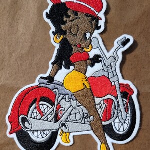 May include: Embroidered patch of a cartoon woman with black hair and a red hat riding a red motorcycle. The woman is wearing a red top and yellow shorts.