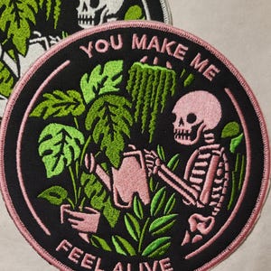May include: A black and pink embroidered patch with a skeleton watering plants. The patch has the text "You Make Me Feel Alive" around the edge.