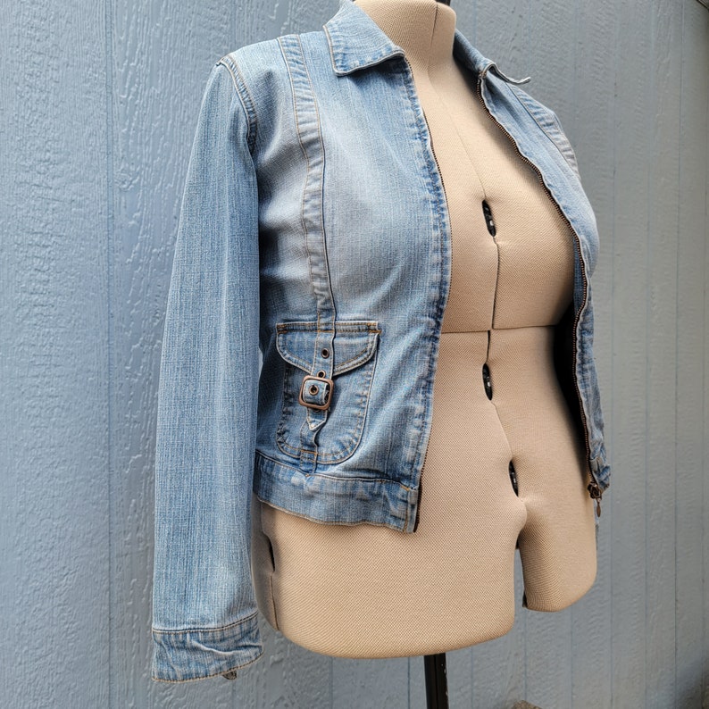 May include: A light blue denim jacket with a zipper closure and a single pocket on the left side. The jacket has a slightly cropped length.