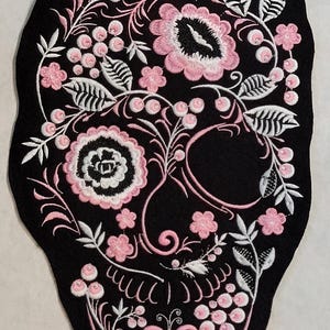 May include: A black fabric patch shaped like a skull, featuring intricate embroidery in shades of pink and white. The design includes floral motifs, leaves, and small berries arranged to form the facial features of the skull on a dark background.
