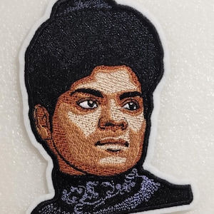 May include: An embroidered patch featuring a portrait of a person with dark skin and a black afro hairstyle. The person wears a black high-neck garment with a subtle purple floral pattern around the collar. The patch has a white border outline.