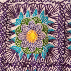 May include: Close-up of white fabric with detailed embroidery. The design showcases a central flower with a yellow centre, encircled by purple, green, and blue elements. The edges are framed with a repeating purple pattern.
