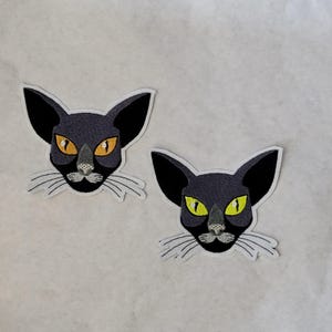 May include: Two embroidered patches featuring a stylized black cat face with large ears and yellow eyes. The cat has a gray nose and white whiskers. The patches are outlined in white and are on a light background.