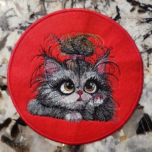 May include: A round red felt patch with an embroidered image of a gray cat with a black nose and big brown eyes. The cat has its hair tied up in a bun and is wearing a pink ribbon. The cat is looking at the viewer and has its paw raised.