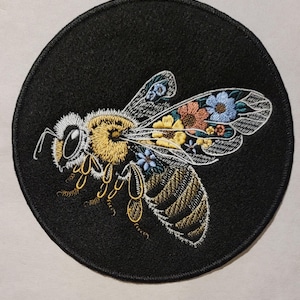 May include: A black embroidered patch with a white bee with yellow and gold accents. The bee is surrounded by colorful flowers.