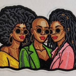 May include: An embroidered patch featuring three figures wearing round sunglasses. The left figure has a large afro and yellow top, the center figure is bald and wears a green blazer, and the right figure has long dark braids and a pink blazer.