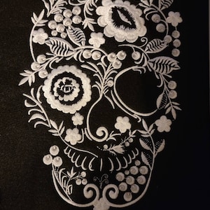 May include: White embroidered floral design on a black background, resembling a sugar skull.
