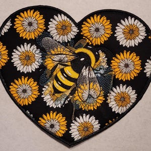 May include: Heart-shaped patch featuring a detailed bumblebee design with yellow and black stripes, surrounded by yellow and white daisies on a black background. The patch is an embroidered design.