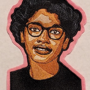 May include: Embroidered patch featuring a portrait of a person with dark curly hair, glasses, and a black shirt. The patch has a pink border and is made with detailed stitching in shades of brown, black, and tan.