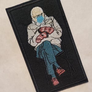 May include: Embroidered patch featuring a person wearing a white coat, blue jeans, and a blue face mask. The person is sitting on a chair and holding a pair of red mittens.