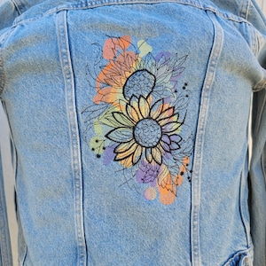 May include: A denim jacket with a colorful embroidered design of two sunflowers with leaves and stems. The embroidery is in shades of orange, yellow, green, purple, and blue.