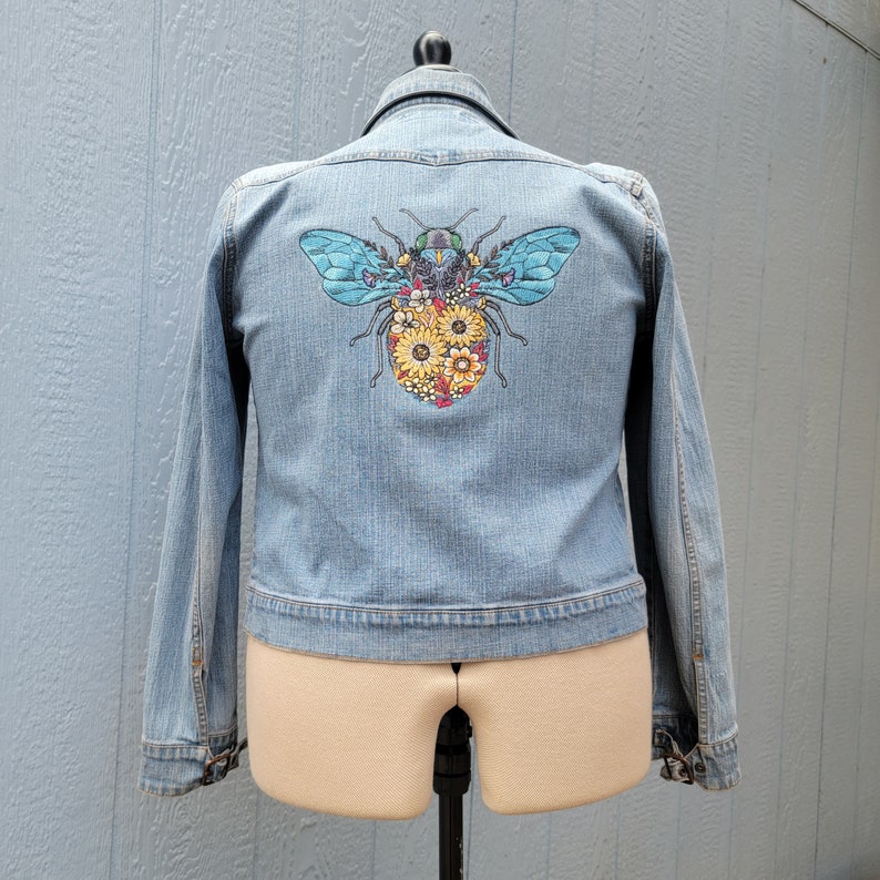 May include: A light blue denim jacket with a floral bee embroidered on the back.