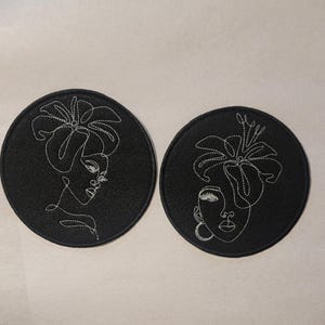 May include: Two black felt circles with white embroidered line art designs of women's faces with floral accents.