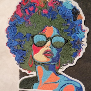May include: Embroidered patch featuring a colorful portrait of a person with an afro hairstyle wearing sunglasses. The portrait is done in a pop art style with bold colors and outlines.