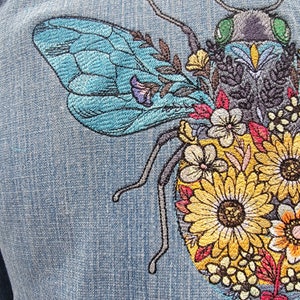 May include: Embroidered denim patch featuring a blue and black bee with a green head surrounded by yellow, red, and white flowers.