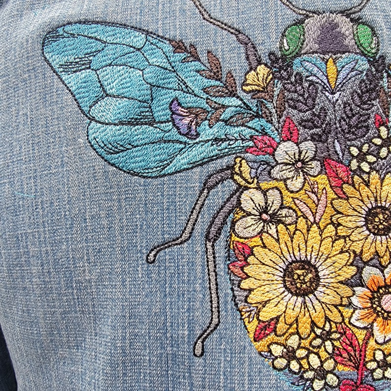 May include: Embroidered denim patch featuring a blue and black bee with a green head surrounded by yellow, red, and white flowers.