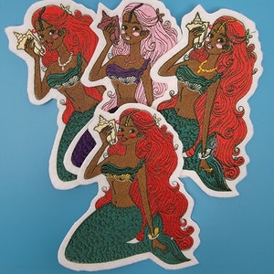 May include: Three embroidered iron-on patches featuring mermaids with long flowing hair, holding seashells. The mermaids are wearing seashell bras and have green tails. The patches are on a blue background.