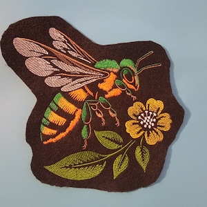 May include: Embroidered patch featuring a bee with orange and green stripes, pink wings, and a yellow flower with a pink center. The bee is flying towards the flower, which is on a brown background.