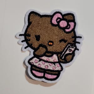 May include: An embroidered iron-on patch featuring a brown cat character with a pink bow, wearing a light pink dress with darker pink polka dots. The character is holding a small white rectangular object, has black whiskers, and a small orange nose.