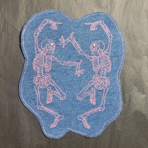May include: Embroidered denim patch featuring two pink skeletons dancing. The patch has a blue denim background with a light blue stitched border. The skeletons are in a playful pose, with arms raised and fingers pointing.