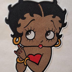 May include: Embroidered patch featuring a cartoon character with black hair, brown skin, and a red heart on her chest. She is wearing gold earrings and a gold bracelet.