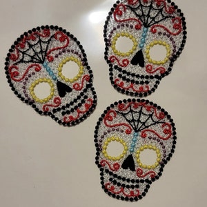 May include: Three embroidered sugar skull patches. Each skull features a white base with yellow eye sockets, a black nose, and a black and red decorative border. The design includes a black spiderweb and a blue vertical line.
