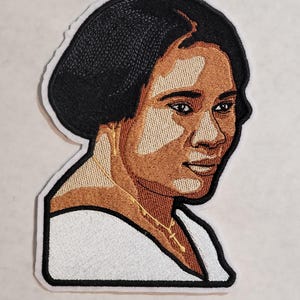 May include: An embroidered patch featuring a portrait of a person with dark hair, brown skin, and vitiligo. The person wears a white top and a thin gold necklace with a small cross pendant. The patch has a thick black border and a white outer edge.