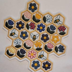 May include: Embroidered patch in a honeycomb design, featuring various colorful flowers and bees. The patch has a white background with gold-colored stitching outlining the honeycomb cells and the floral and bee details. The flowers are in shades of blue, pink, yellow, and white.