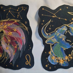 May include: Two embroidered patches depicting the zodiac signs Leo and Taurus. The patches are black with gold embroidery and feature the constellations for each sign. The Leo patch features a lion's head with a mane of gold and red, while the Taurus patch features a bull's head with a blue and green body.