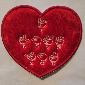 May include: Red felt heart-shaped patch with white embroidered letters spelling "I LOVE YOU" in American Sign Language.