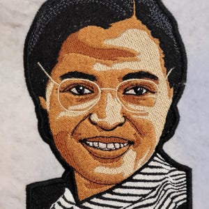 May include: An embroidered patch featuring a portrait of a person with dark hair, wearing thin-rimmed glasses and a black and white striped shirt. The design is rendered in shades of brown, tan, and black thread with a thick black stitched border.