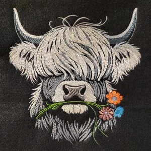 May include: Embroidered design of a white Highland cow with long, shaggy hair, holding a bouquet of pink, orange, and blue flowers in its mouth.