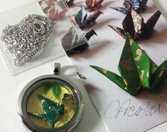 Può includere: Una collana con un medaglione color argento con una piccola gru di origami all'interno, accanto a una catena d'argento. Sullo sfondo una carta con gru di origami. La gru di origami all'interno del medaglione è verde acqua e gialla.