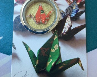 Può includere: Una collana con pendente in argento con una gru di carta all'interno di un medaglione rotondo di vetro. La gru è rossa e dorata. Una gru origami verde con accenti dorati è sulla carta sotto il pendente. La carta ha il nome scritto a mano "Oresta".