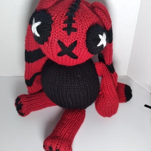 May include: A red and black knitted bunny rabbit plush toy with stitched-on eyes and a stitched-on mouth. The bunny has long ears and a black body.