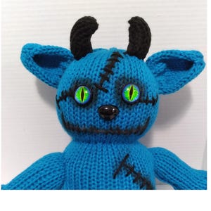 Puede incluir: Un juguete de punto azul con cuernos negros, costuras cosidas y ojos verdes. El juguete tiene una nariz negra y una boca cosida.