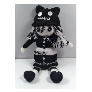 Pode incluir: Uma boneca de malha preta e branca com o rosto costurado, usando um chapéu preto com orelhas de gato e um vestido listrado preto e branco. A boneca tem olhos de botão e uma boca costurada.