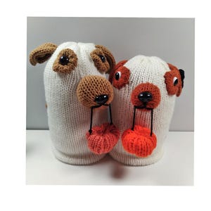 Pode incluir: Dois gorros brancos de malha com rostos de cão. Os chapéus têm orelhas, olhos e narizes castanhos, com pompons laranja pendurados nos narizes. Os chapéus são projetados para se parecerem com cães.