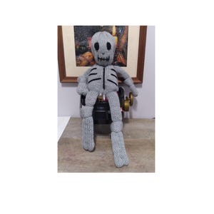 May include: A grey knitted skeleton doll with black eyes and a black mouth sitting on a black chair. The doll has a knitted ribcage and knitted arms and legs.
