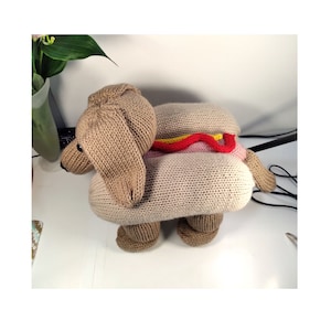 May include: A knitted dachshund dog toy with a hot dog design. The dog is brown and the hot dog is pink with yellow mustard and red ketchup.