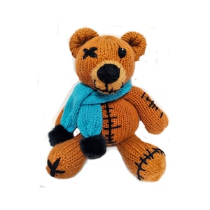 May include: A brown knitted teddy bear wearing a blue scarf with black pom-poms. The bear has a stitched-up mouth and one eye is stitched shut with an X.